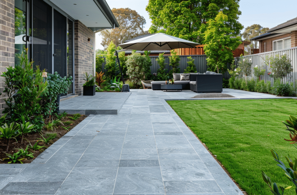 Modern patio paving in a Rowville backyard featuring large grey stone pavers, fresh green lawn, and landscaped garden beds with outdoor seating and umbrella, realistic Melbourne suburban setting.